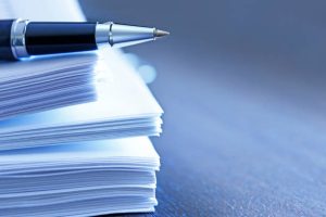 A ballpoint pen rests on top of a stack of documents ready for signing.  The image is photographed using a very shallow depth of field with the focus being on the tip of the pen.